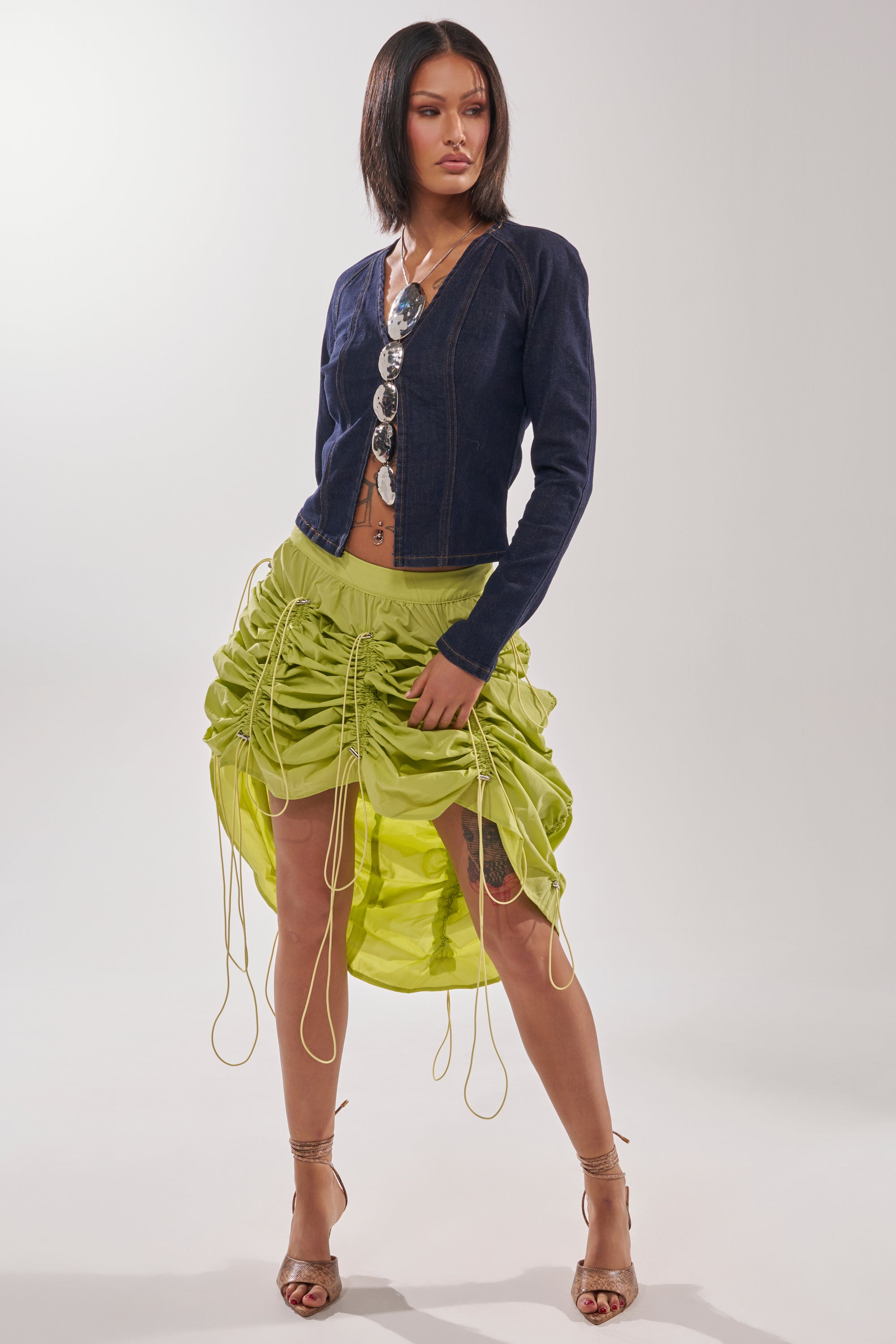 A woman stands confidently in a navy denim jacket, statement necklace, and the BELLA PARACHUTE MAXI SKIRT IN YELLOW with drawstrings. She pairs strappy heels with the look and poses against a plain white background. 9