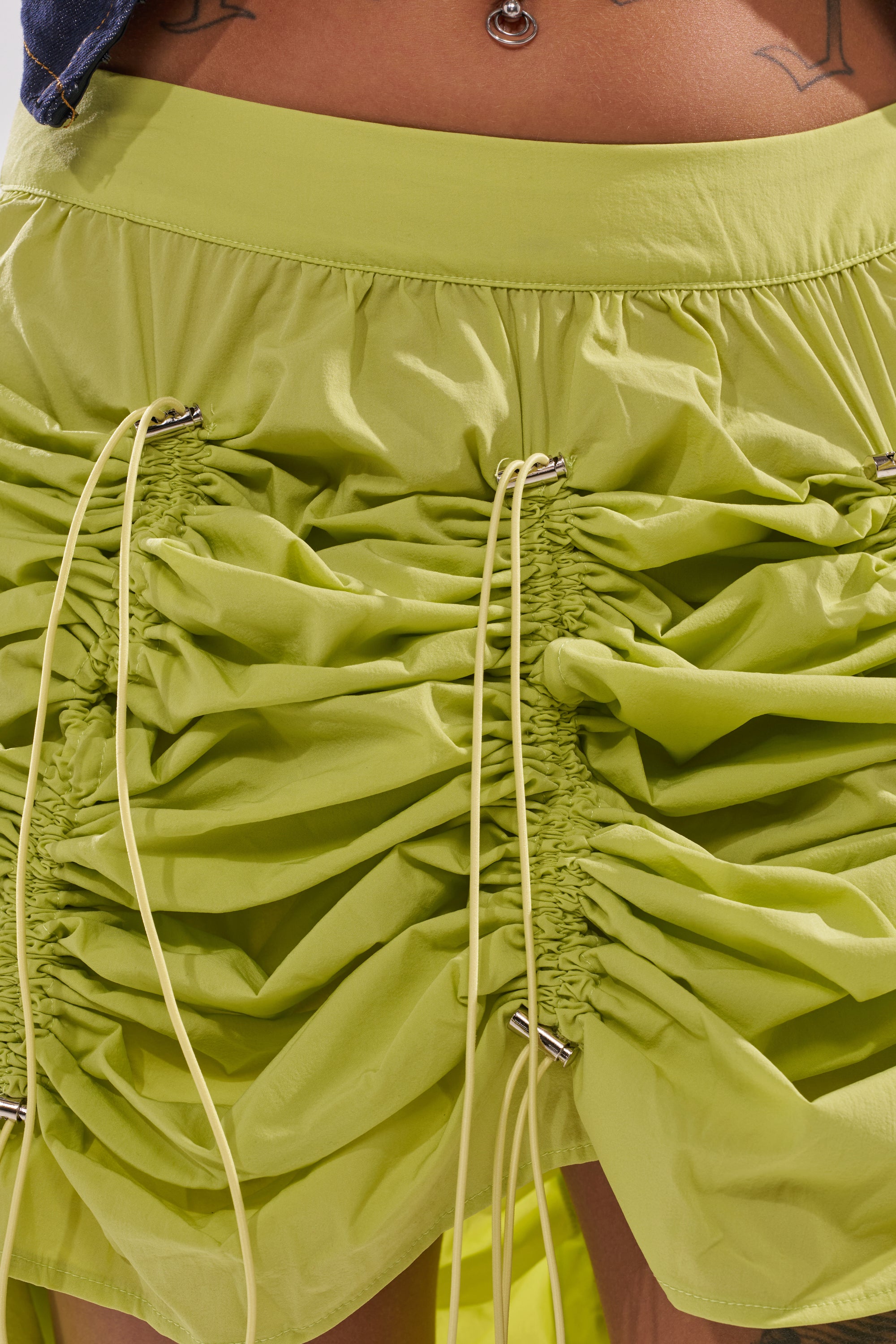 Close-up of the BELLA Parachute Maxi Skirt in yellow, featuring an adjustable drawcord and vertical ruching with gathered drawstrings. The skirt is paired with a visible belly button piercing and a hint of tattoo above the waistband. 12