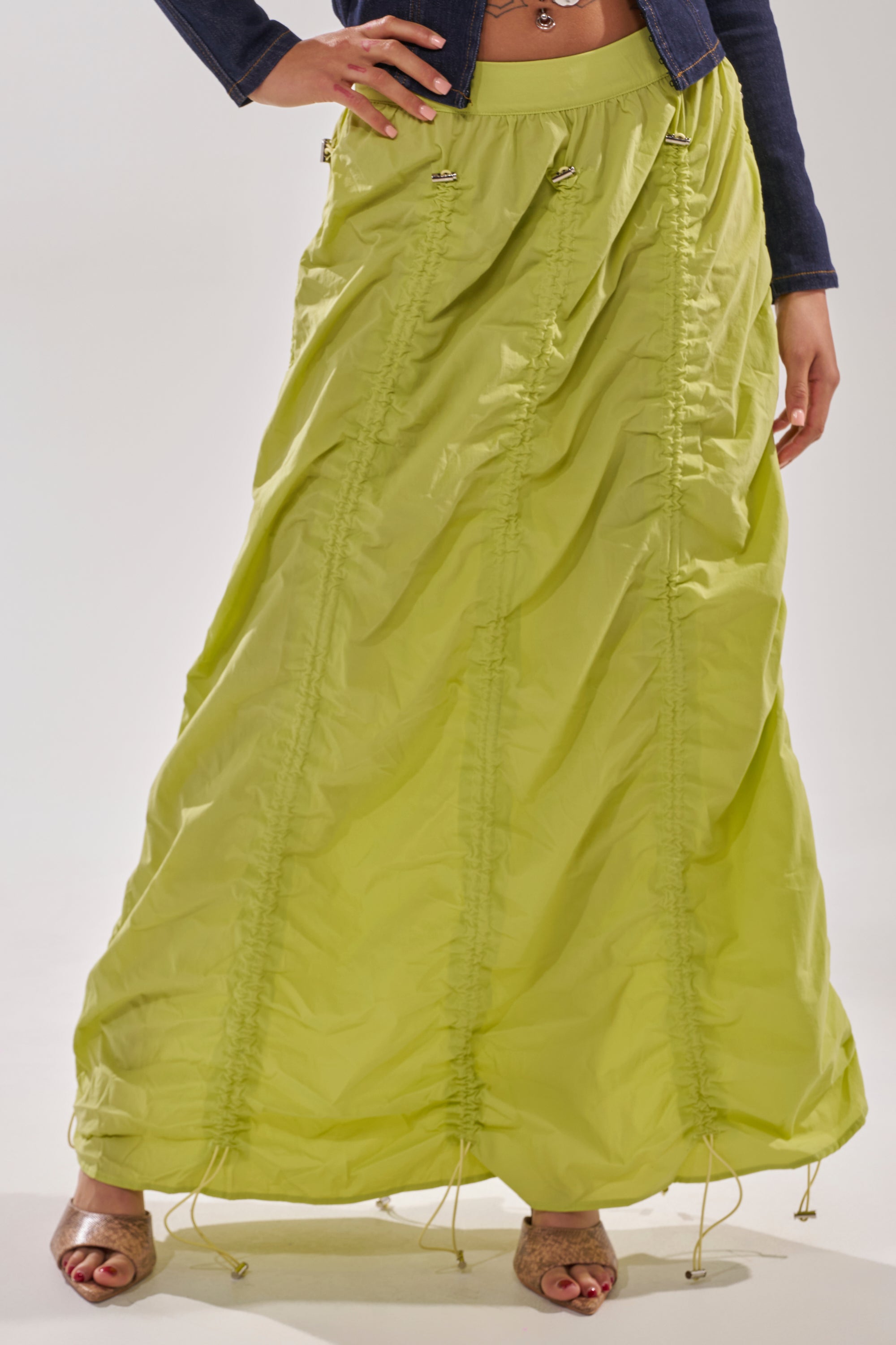 A person stands against a plain light background wearing the BELLA PARACHUTE MAXI SKIRT IN YELLOW with ruching and an adjustable drawcord, styled with a dark cropped jacket and open-toe heels. 2