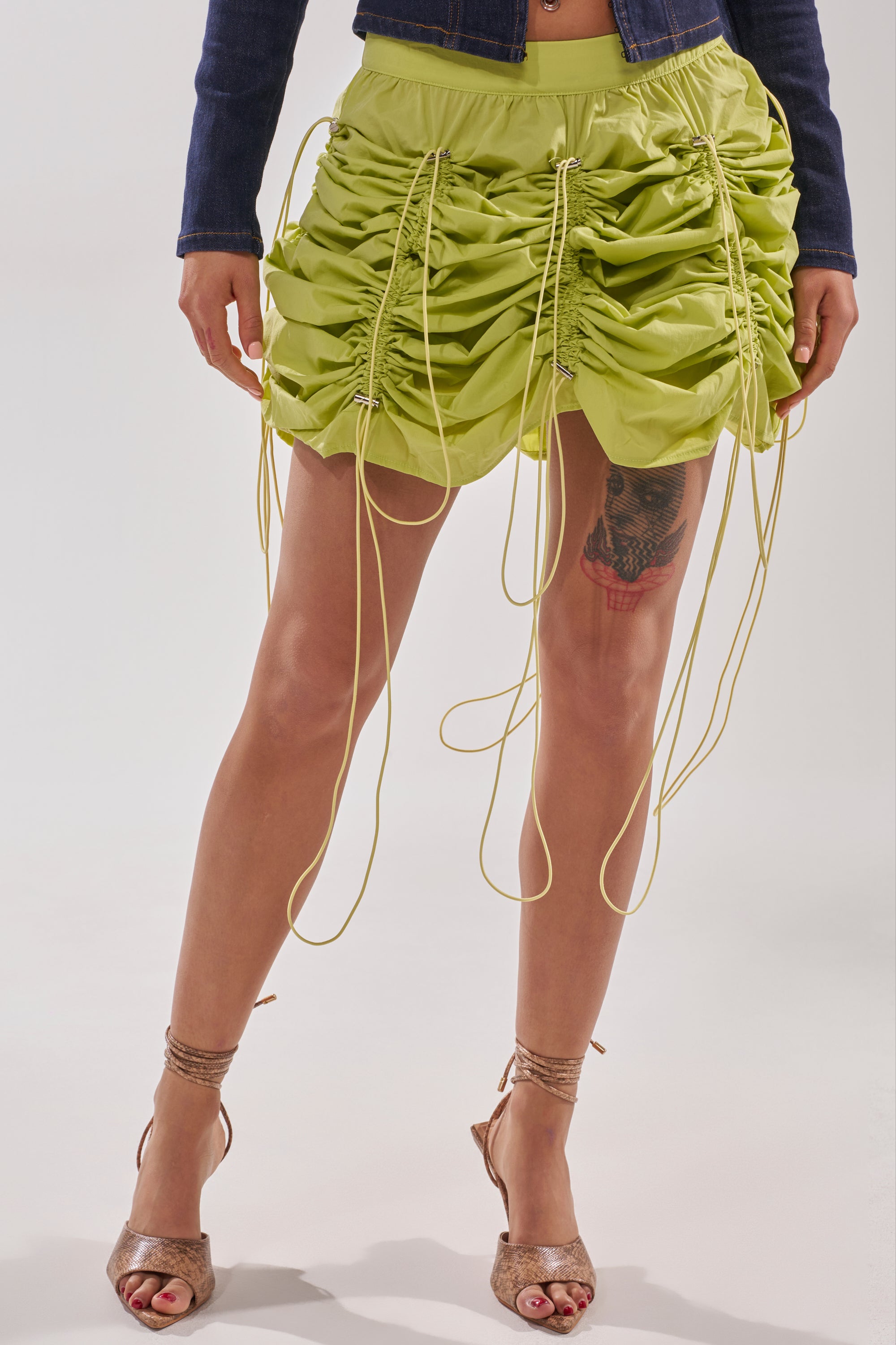 A person wears the BELLA PARACHUTE MAXI SKIRT IN YELLOW with an adjustable drawcord and long ties, styled with a cropped denim jacket and strappy heels. A colorful thigh tattoo on their right leg adds flair to the trendy outfit. 3