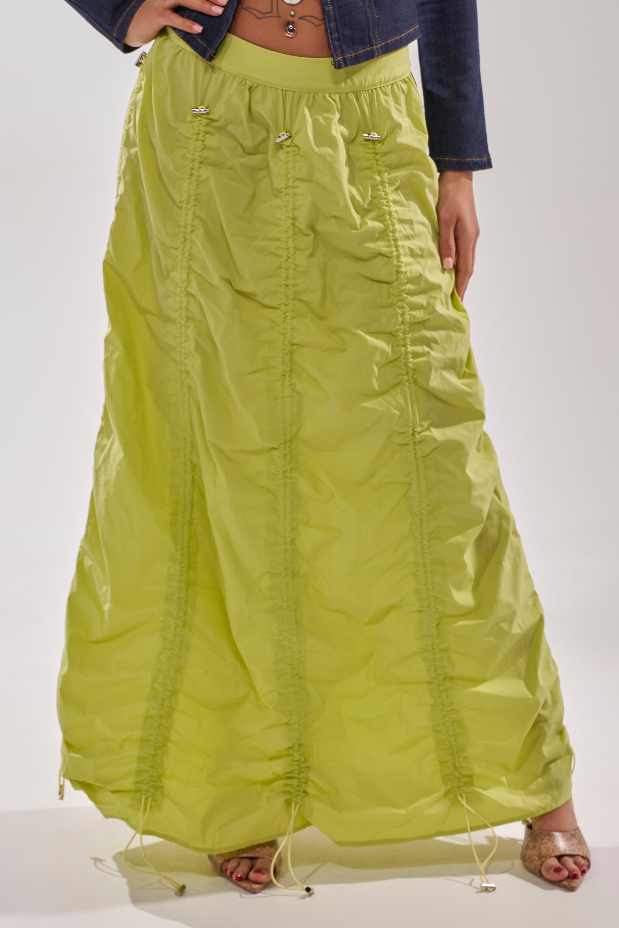Wearing the BELLA PARACHUTE MAXI SKIRT IN YELLOW with an adjustable drawcord and ruched detailing, a dark denim jacket, and open-toe shoes, a person stands against a plain background. 4