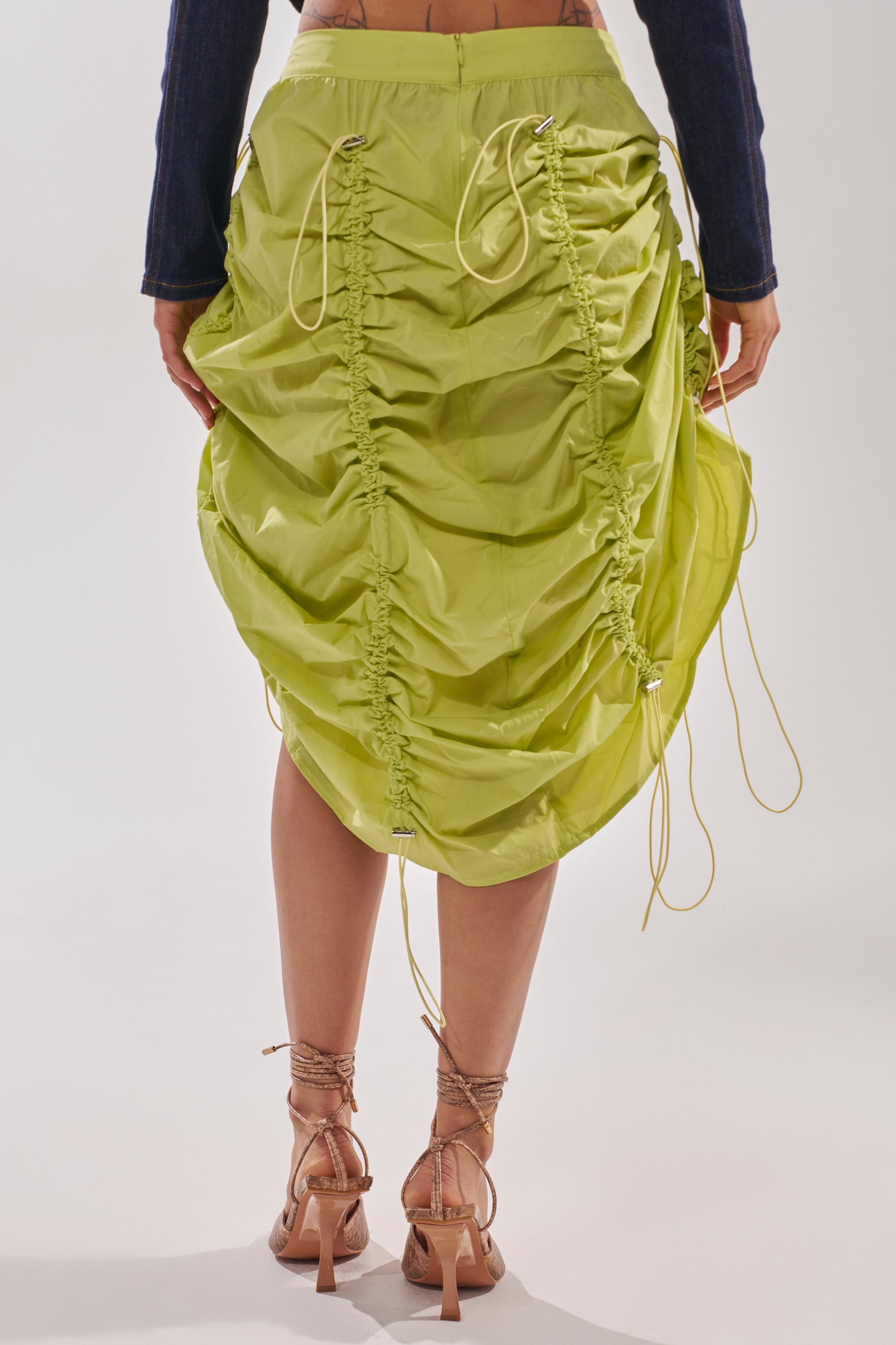 A person stands facing away, wearing the BELLA PARACHUTE MAXI SKIRT IN YELLOW with adjustable drawcord and ruched details, paired with strappy high-heeled sandals. 6