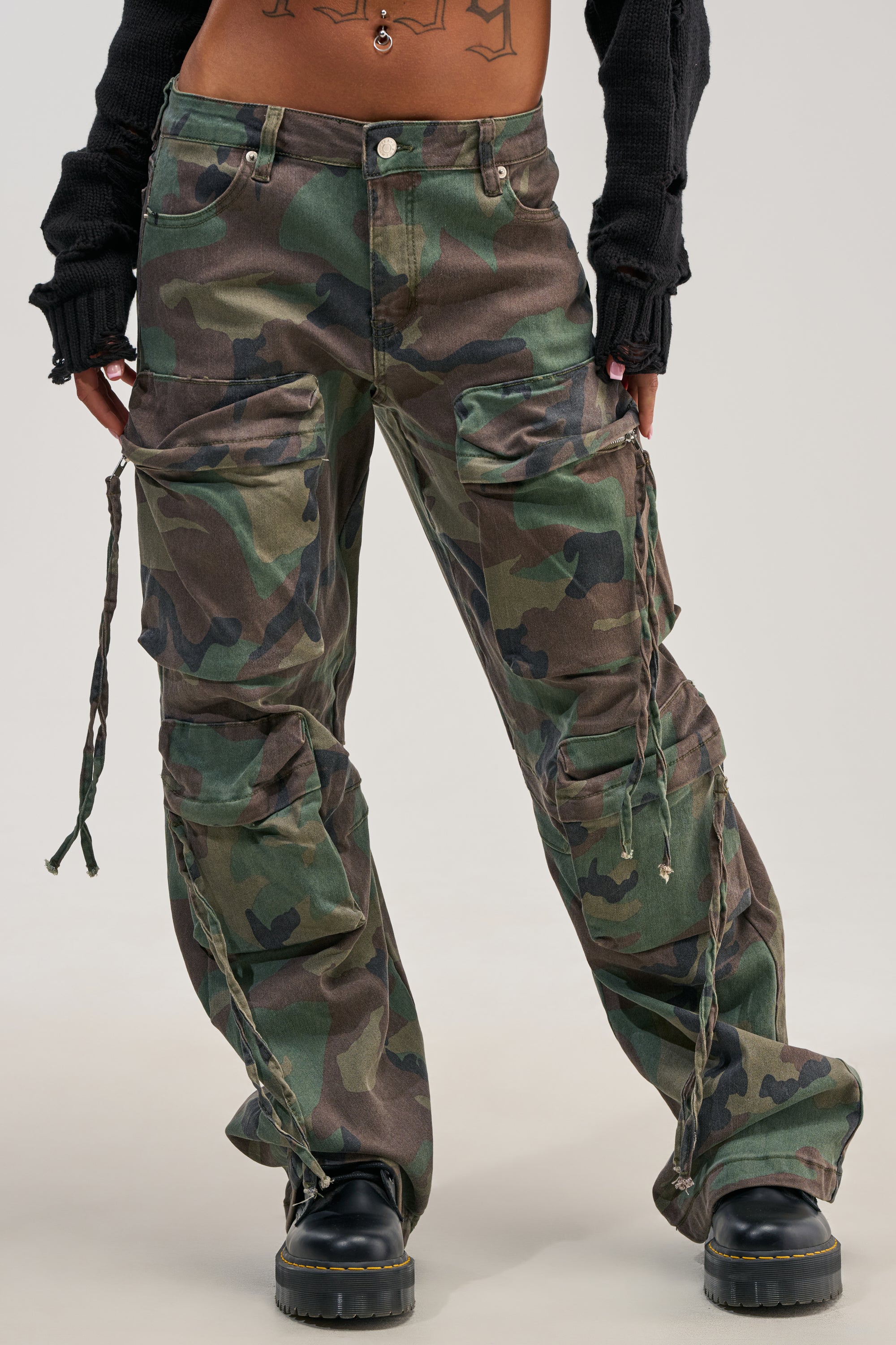 Person wearing the TAKE ME DOWN CAMO CARGO PANT, a loose, streetwear-inspired camo cargo pant featuring large side pockets and drawstring details, styled with black boots, a cropped black sweater, and sporting a visible tattoo on their torso. The background is plain and light-colored.