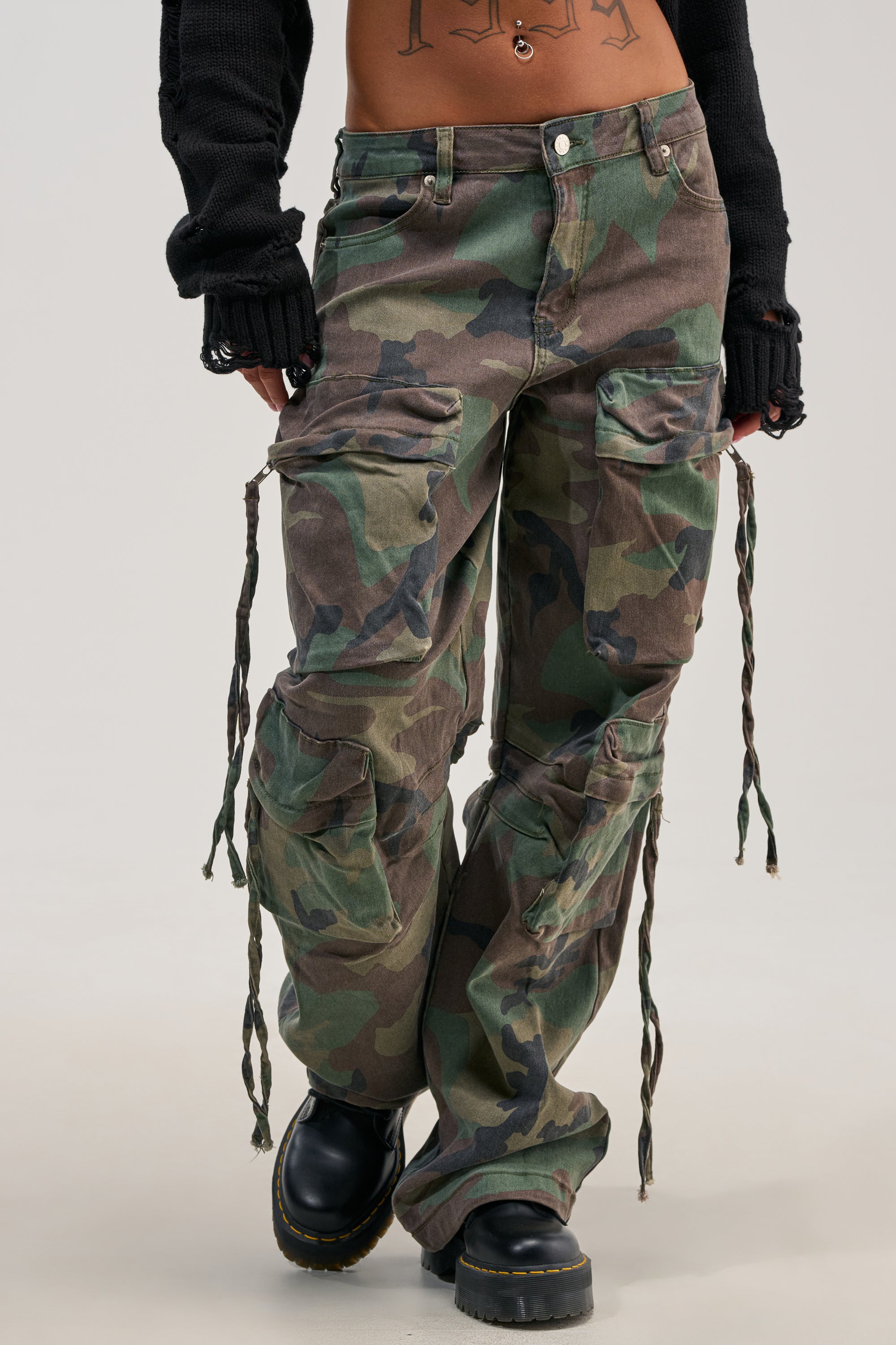 Wearing the TAKE ME DOWN CAMO CARGO PANT featuring drawstring details, black shoes, and a black long sleeve top, this person creates a bold streetwear look. Tattoos are visible on their midriff, set against a plain light gray background.