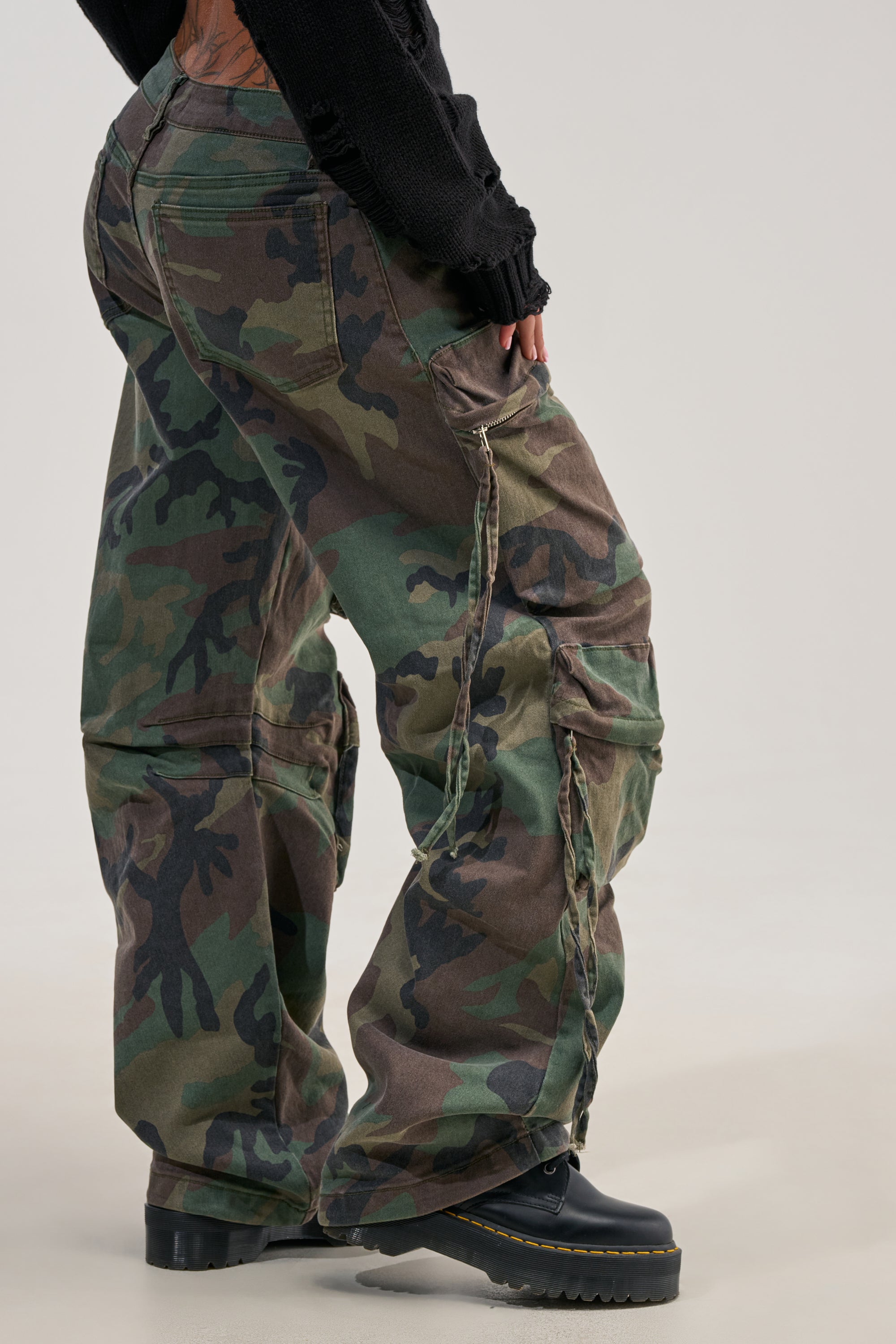 Wearing the TAKE ME DOWN CAMO CARGO PANT styled with black platform shoes and a black long-sleeve top, the person is photographed from behind to highlight the streetwear-inspired fit and design of the cargo pants against a neutral background.