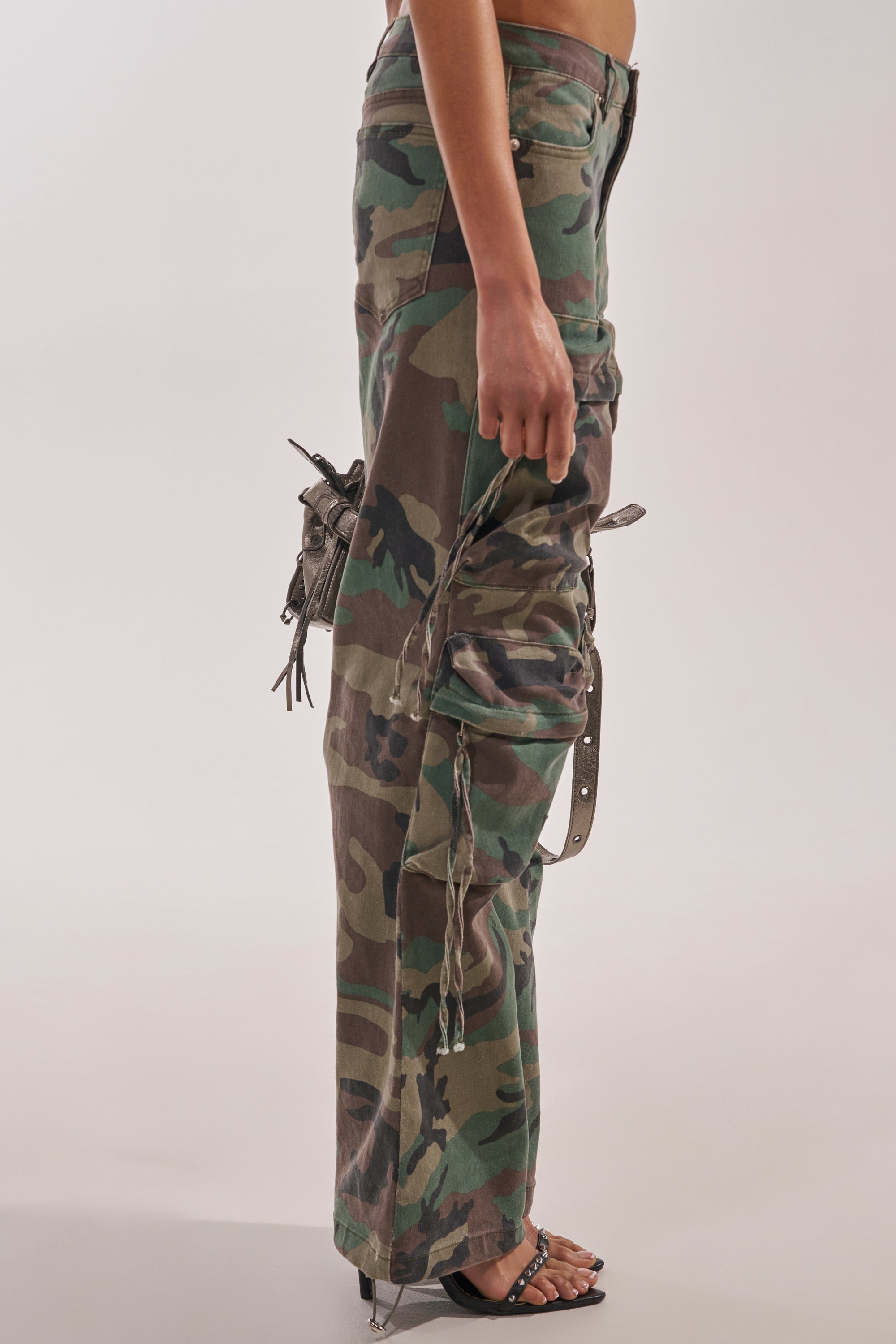 A person in profile wears the TAKE ME DOWN CAMO CARGO PANT, featuring a green camo print, wide-leg silhouette, and drawstrings, paired with black sandals and holding a small bag against a plain light background. 3