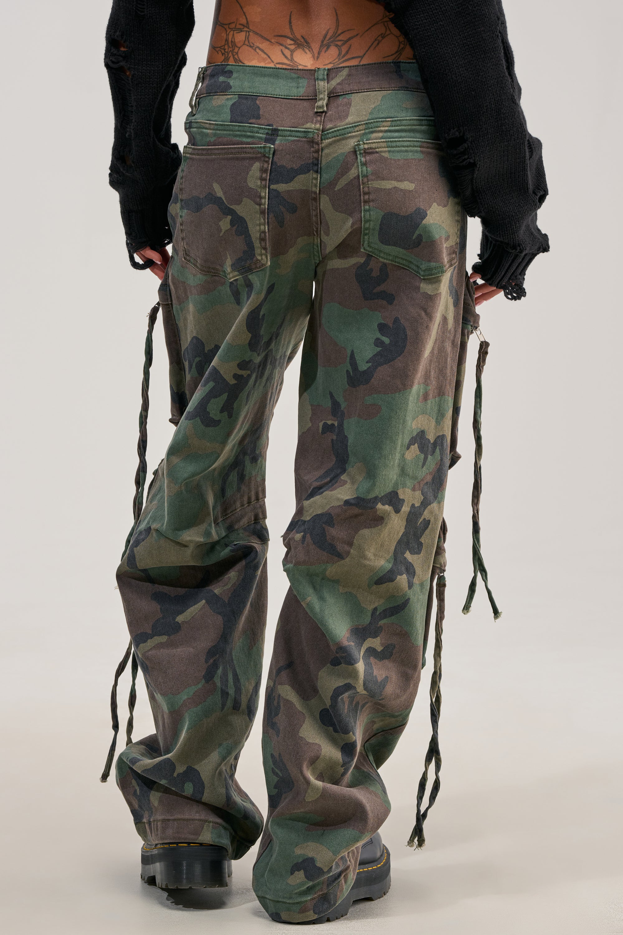 Seen from behind, a person wears the TAKE ME DOWN CAMO CARGO PANT—loose, wide-leg cargo pants with standout streetwear style, featuring ties hanging from the sides—paired with black boots. They also sport a black ripped sweater, with tattoos visible on their lower back.