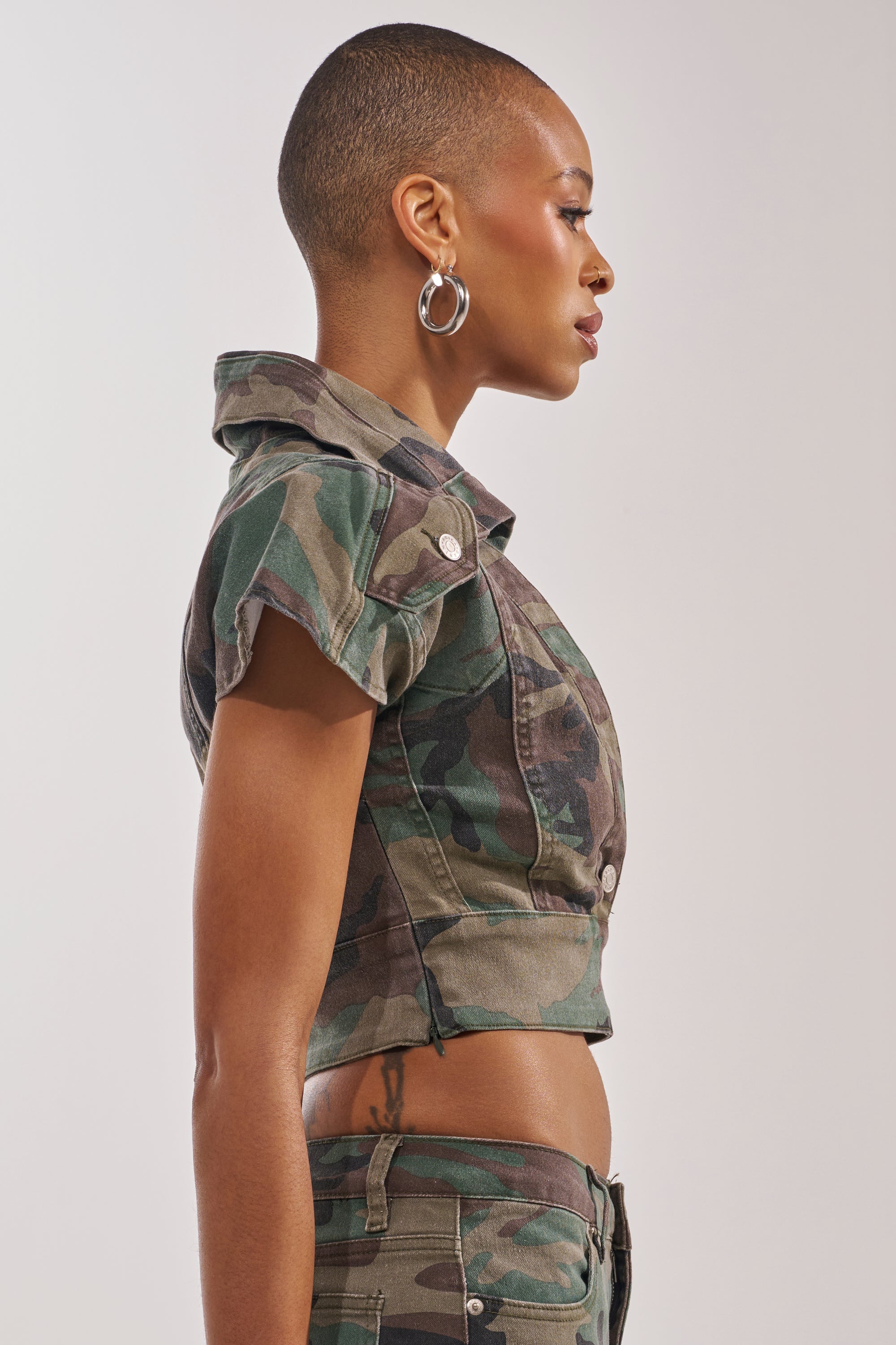 A person with a shaved head and hoop earrings is shown in profile wearing the TAKE ME DOWN CAMO TOP with matching pants against a plain background. 2