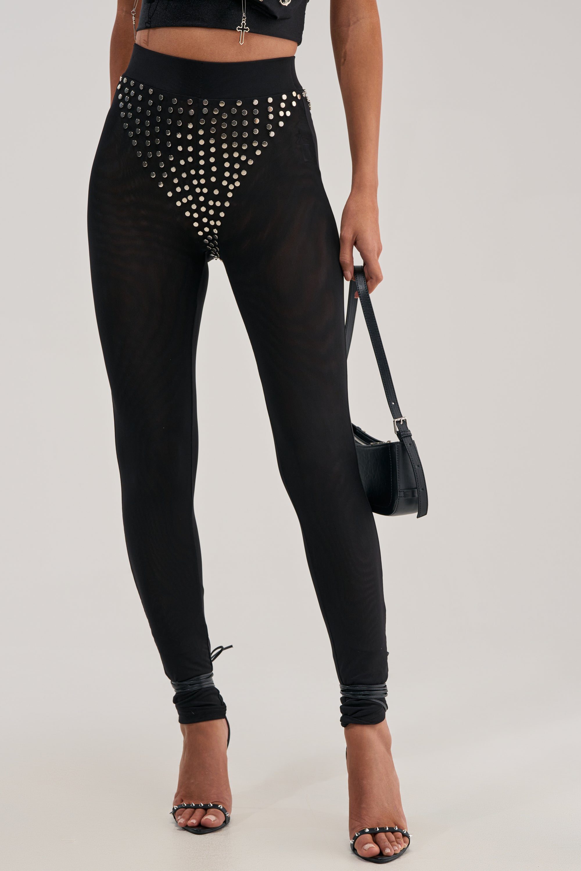A person wearing the COOLEST GIRL ON THE BLOCK Mesh Legging with Studs, featuring silver studs in a triangular pattern, paired with black heeled sandals and holding a small black handbag against a plain background.