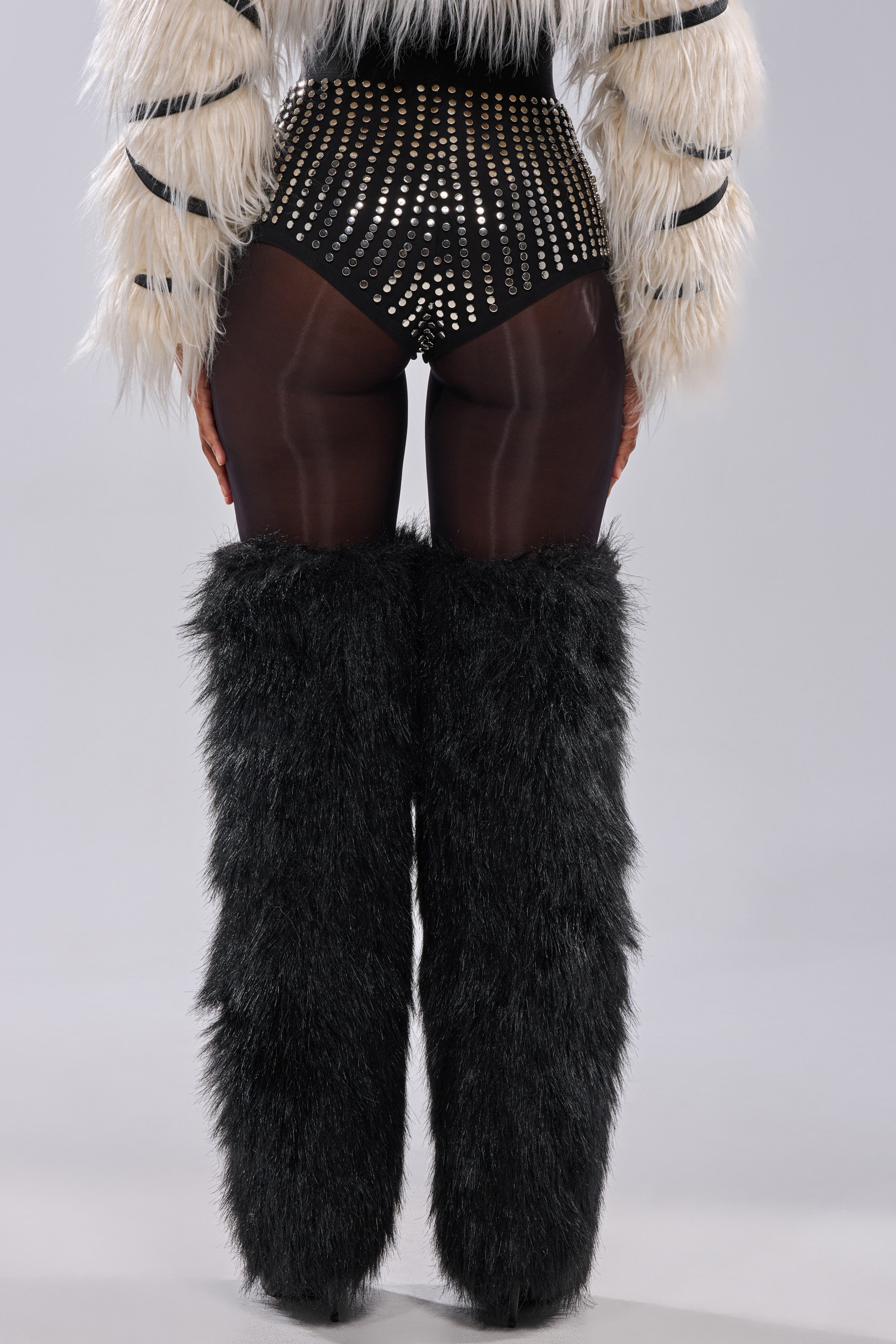 A person stands facing away, wearing the COOLEST GIRL ON THE BLOCK MESH HOT SHORT WITH STUDS, paired with black furry leg warmers, black sheer tights, and a white faux fur jacket with black stripes, against a plain light background. 3