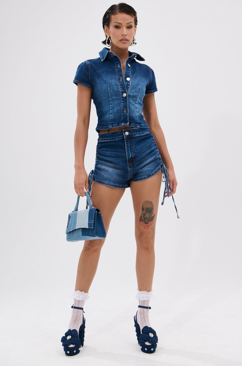 A woman stands confidently in the NEW CANADIAN DENIM SHORT and a denim collared top, holding a matching bag. She pairs blue platform heels with white frilly socks and shows off a skull tattoo on her thigh. 4