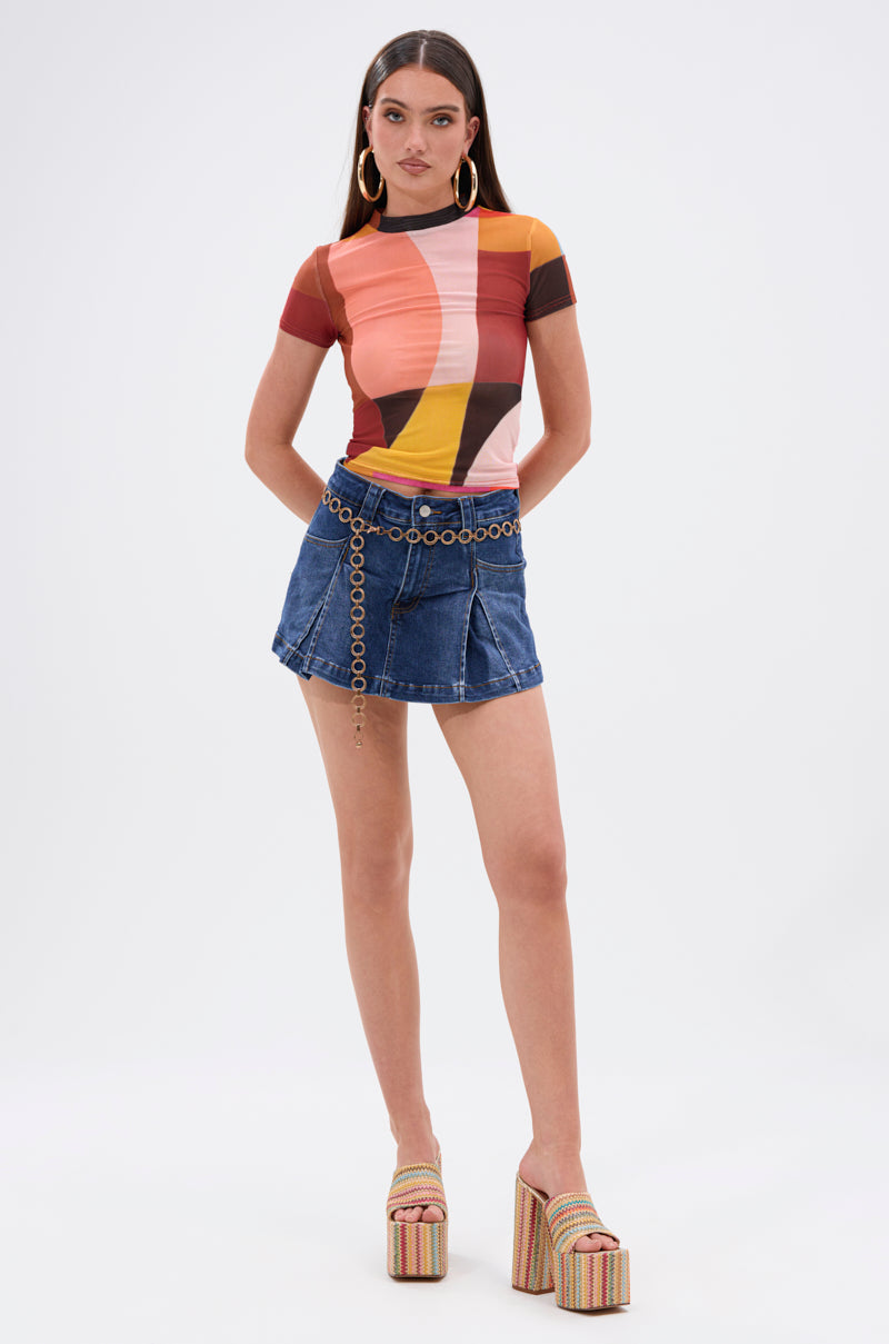 A woman in a colorful geometric tee, JULIE DENIM SKORT with chain belt, gold hoop earrings, and striped platform heels stands against a white background—a bold addition to any casual wardrobe.