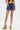 A person wearing a colorful sleeveless top, the JULIE DENIM SKORT with a circular ring belt, and high platform sandals with multicolored stripes, poses with one hand in a pocket against a plain white background.