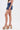 A person standing sideways in a sleeveless top, JULIE DENIM SKORT in indigo wash, and colorful striped platform sandals against a plain white background—perfect for a casual wardrobe.
