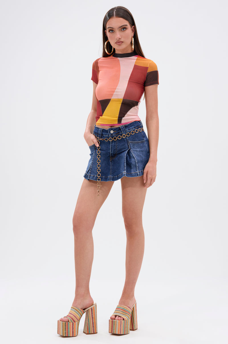 A woman models the JULIE DENIM SKORT in indigo wash with a chain belt, paired with a vibrant geometric top, large hoop earrings, and multicolored platform heels—an effortlessly chic look against a plain white background.