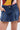 Wearing the JULIE DENIM SKORT in indigo wash with front pockets and a vibrant, geometric-patterned top—one hand tucked into the pocket—for a relaxed, casual look.