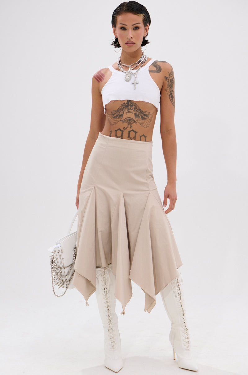 A person with tattoos wears a white sleeveless crop top, the beige HANNAH MIDI SKIRT with handkerchief pleats, and white knee-high lace-up boots. They accessorize with layered necklaces and a white bag, posing against a plain white background. 0