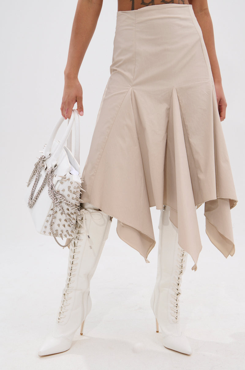 Wearing the HANNAH MIDI SKIRT in high-waist beige with a handkerchief hem, paired with white lace-up high-heeled boots, she holds a white handbag adorned with silver studs and chains against a plain white background. 2
