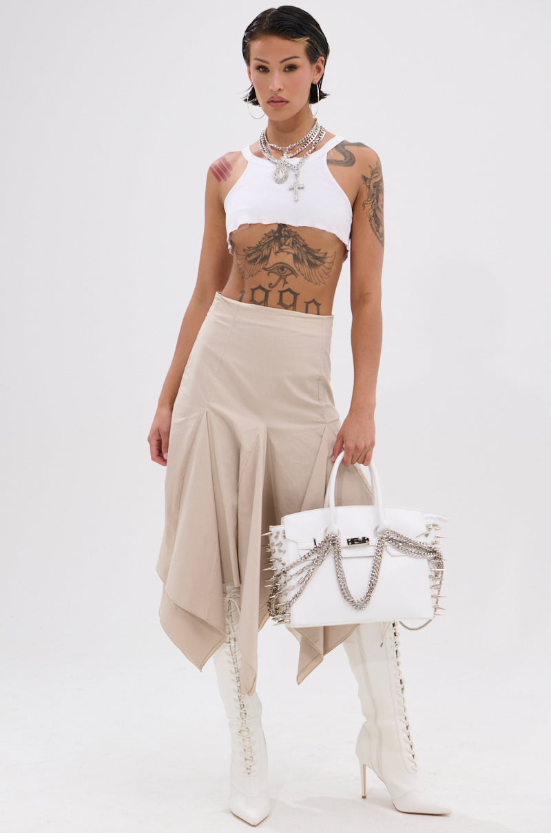 A tattooed woman wears a white crop top, the high-waist HANNAH MIDI SKIRT with handkerchief pleats in beige, white heeled boots, and holds a white chain-detail handbag. Layered necklaces complete the look against a plain white background. 5