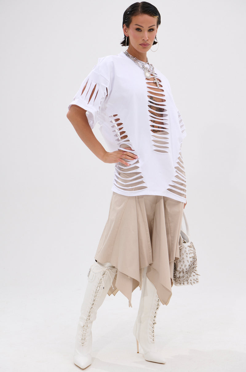 A woman poses confidently in the HANNAH MIDI SKIRT, a high-waist beige midi with handkerchief pleats, styled with a white oversized shredded T-shirt, white lace-up knee-high boots, chunky necklace, and studded handbag. 8