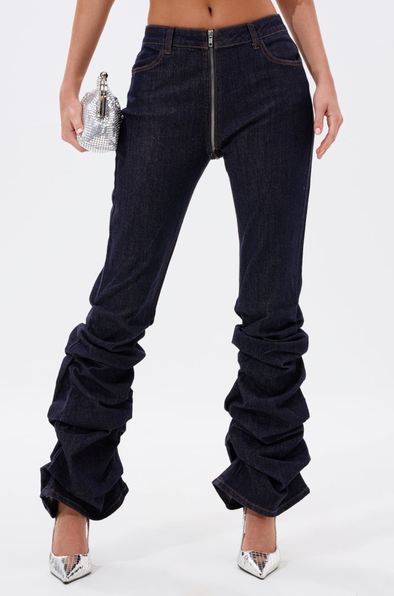 A person wears the GET CRAZY RUCHED DENIM PANT in dark-wash with silver pointed-toe heels, holding a sparkly silver clutch. The waist-down image is set against a plain white background. 2