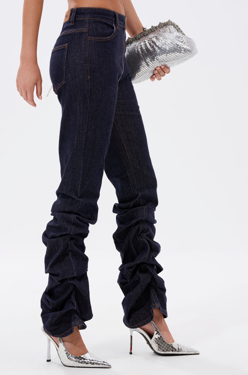 A person wearing the GET CRAZY RUCHED DENIM PANT in dark blue and silver high heels holds a shiny silver textured clutch bag against a plain white background. 3