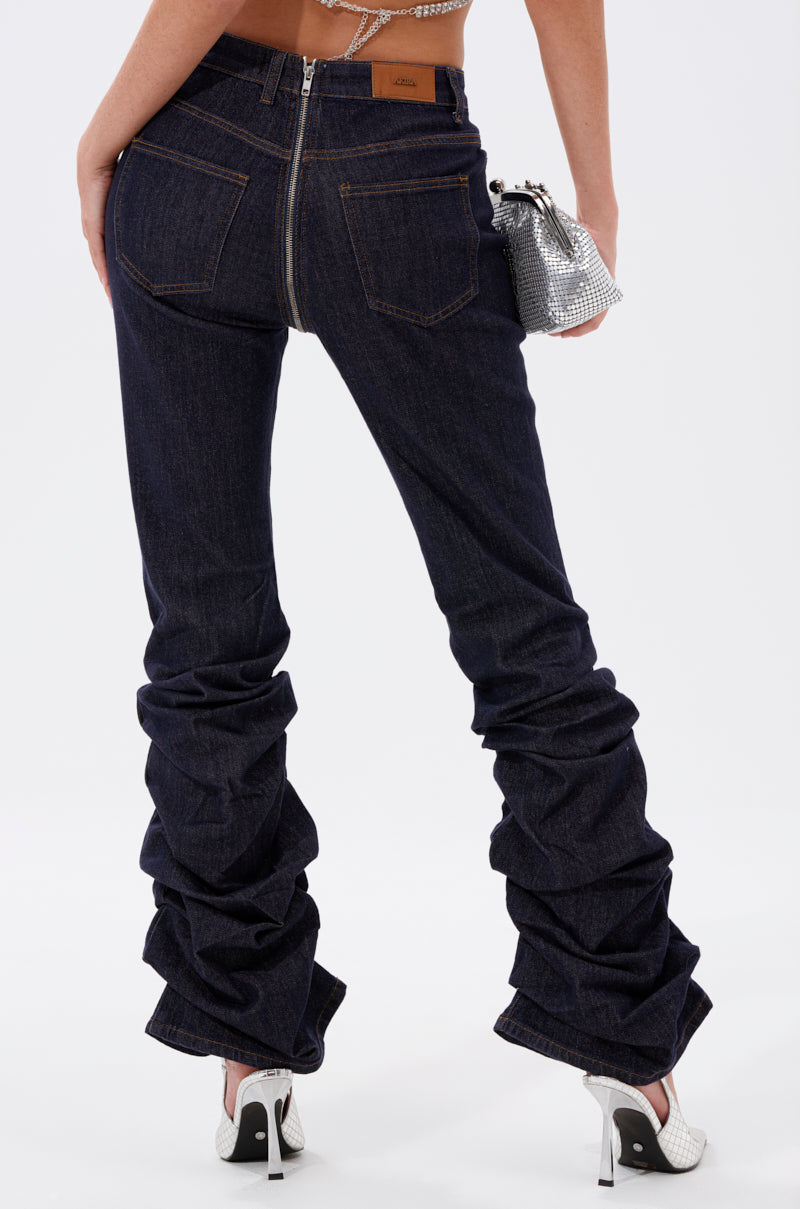 A person is shown from the back against a plain background wearing GET CRAZY RUCHED DENIM PANT in dark blue with a visible back zipper, paired with white high heels and holding a small silver textured bag. 4