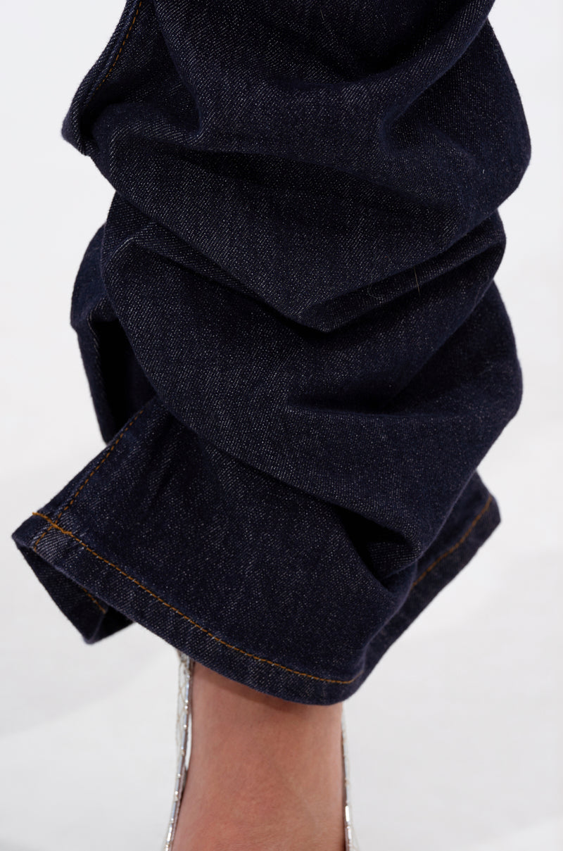 A close-up of a leg wearing the GET CRAZY RUCHED DENIM PANT in dark-wash, cuffed and ruched at the ankle with visible orange stitching, paired with a clear high-heeled shoe. 7
