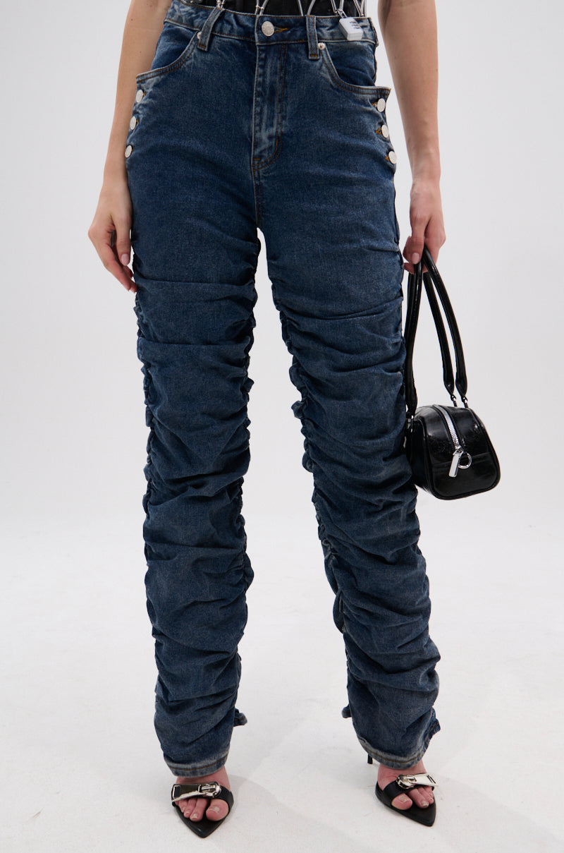 Wearing the VERONICA RUCHED DENIM high-rise, vintage-wash jeans with metal buttons, black heeled sandals, and holding a small black handbag, a person stands against a plain white background.