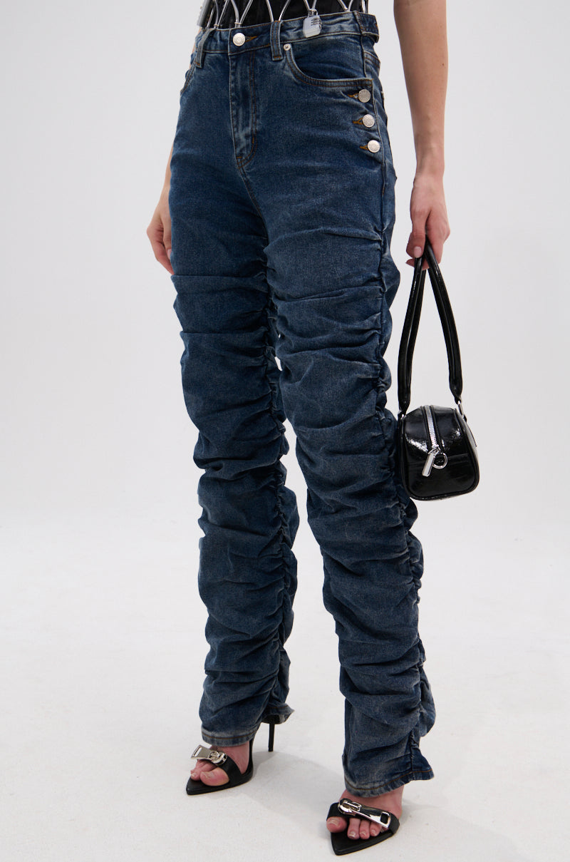 Wearing VERONICA RUCHED DENIM jeans with side metal buttons, black heeled sandals, and a small black handbag against a plain light background.