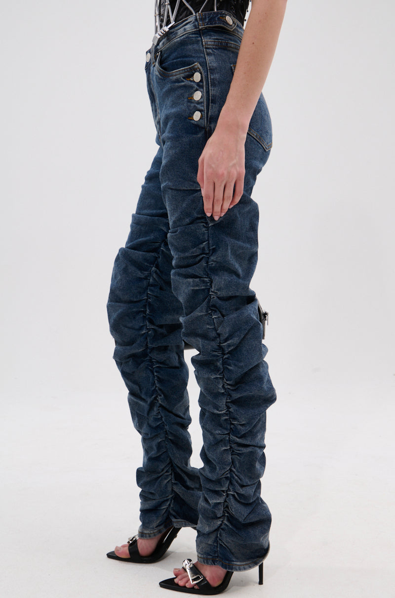 Wearing the VERONICA RUCHED DENIM, featuring a high waist, vintage wash, sculptural silhouette, gathered ruching along the legs, prominent buttons, and paired with black high-heeled sandals.