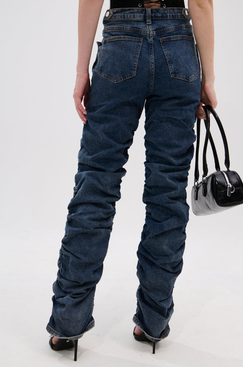 A person faces away, wearing VERONICA RUCHED DENIM high-rise jeans with black high heels and holding a small black handbag against a light, plain background.