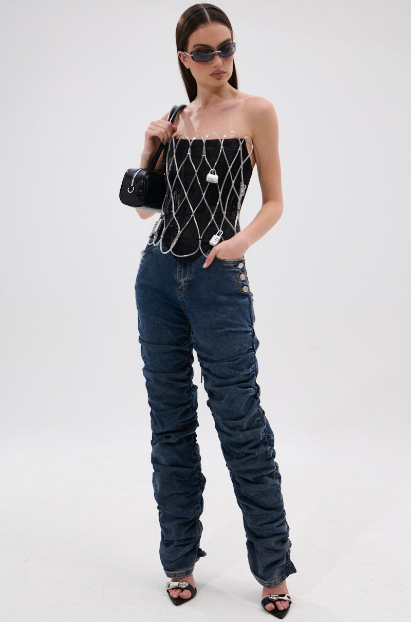 A woman poses against a plain background in dark sunglasses, a black corset with white net overlay, VERONICA RUCHED DENIM jeans with side buttons, black heeled sandals, and a small black handbag.
