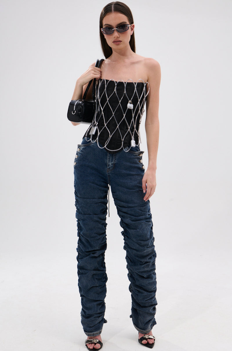 A woman stands against a white background wearing sunglasses, a black strapless top with a chain overlay, black heeled sandals, a small black shoulder handbag, and VERONICA RUCHED DENIM jeans.