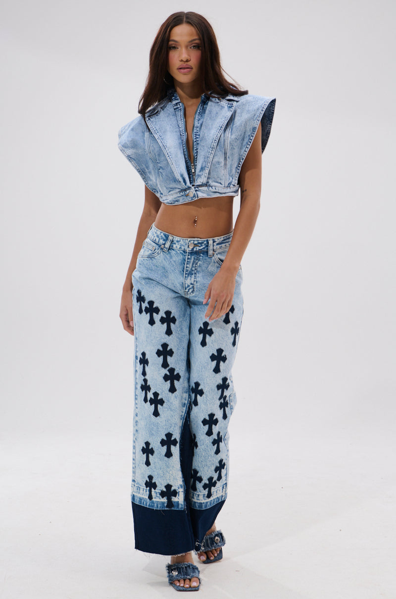 Wearing the CROSSED OUT WIDE LEG DENIM in vintage light-wash with black cross patterns, a woman pairs it with a sleeveless, cropped denim top and denim sandals, walking confidently against a plain white background.