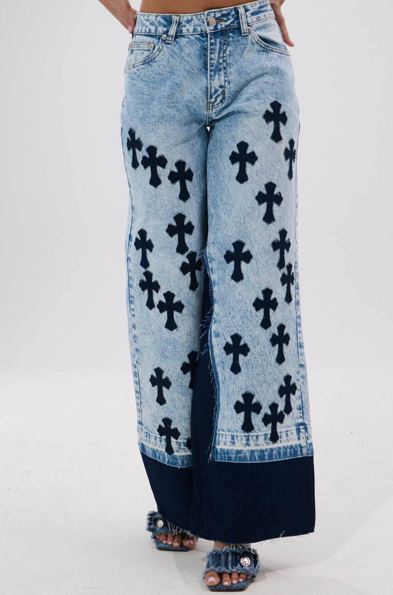Wearing CROSSED OUT WIDE LEG DENIM, featuring vintage light-wash, wide-leg jeans with dark blue cross patterns and a solid blue hem, paired with open-toe fringed sandals. Photo is cropped above the waist and below the ankles.
