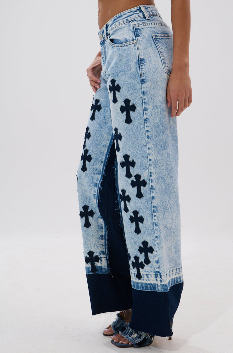Wearing the CROSSED OUT WIDE LEG DENIM—vintage light-wash, high-rise jeans with black cross patterns, a high front slit, raw hems, wide legs, and dark blue panels at the bottom—stands against a plain white background.