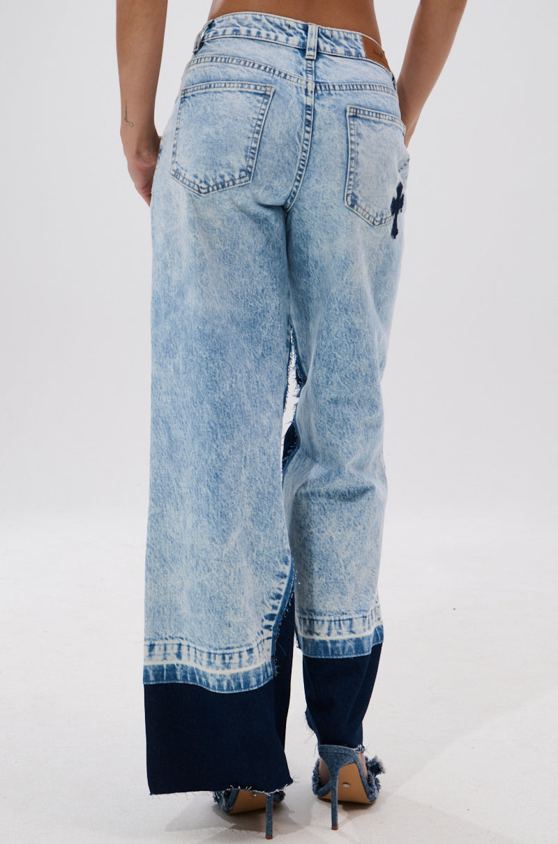 A person seen from the back wears CROSSED OUT WIDE LEG DENIM: vintage light-wash, high-rise jeans with dark blue cuff panels, a black cross patch on the right pocket, wide legs, and frayed hems.
