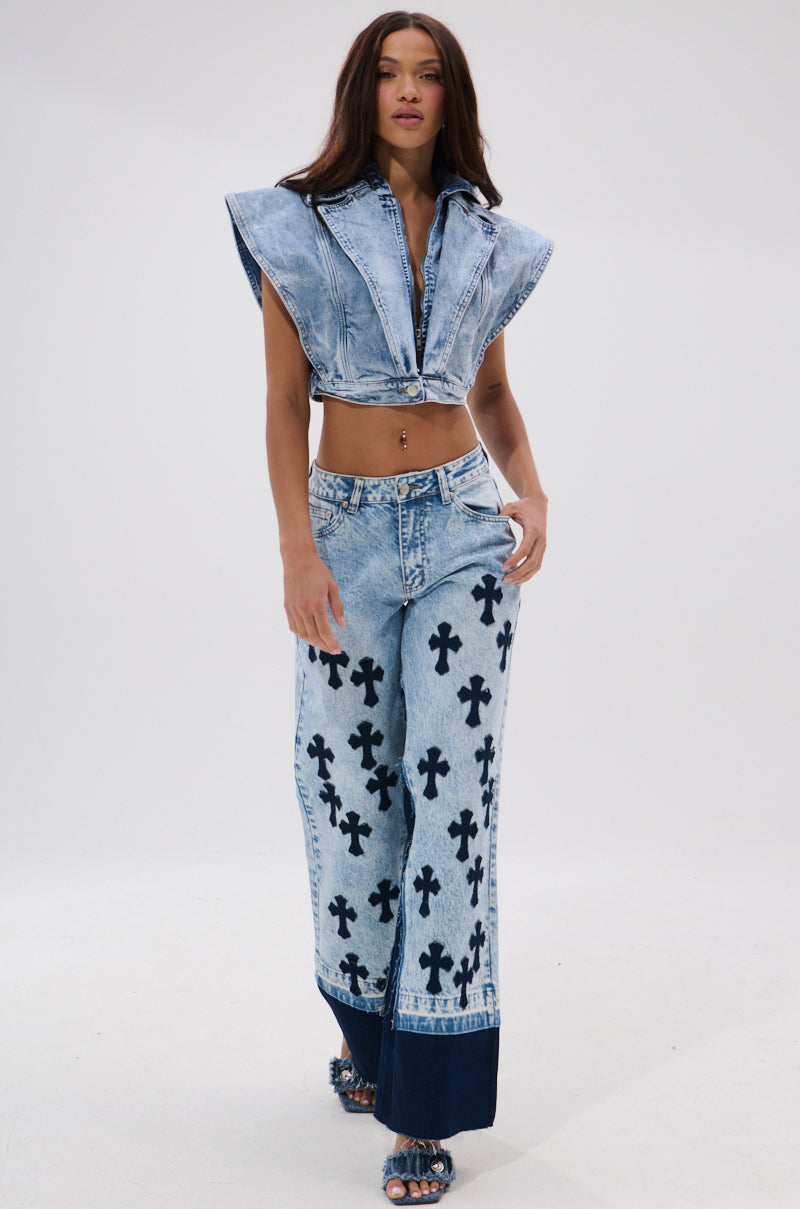 A woman wears the CROSSED OUT WIDE LEG DENIM and a matching vintage light-wash denim crop top, both with black cross patterns, standing confidently against a plain white background.