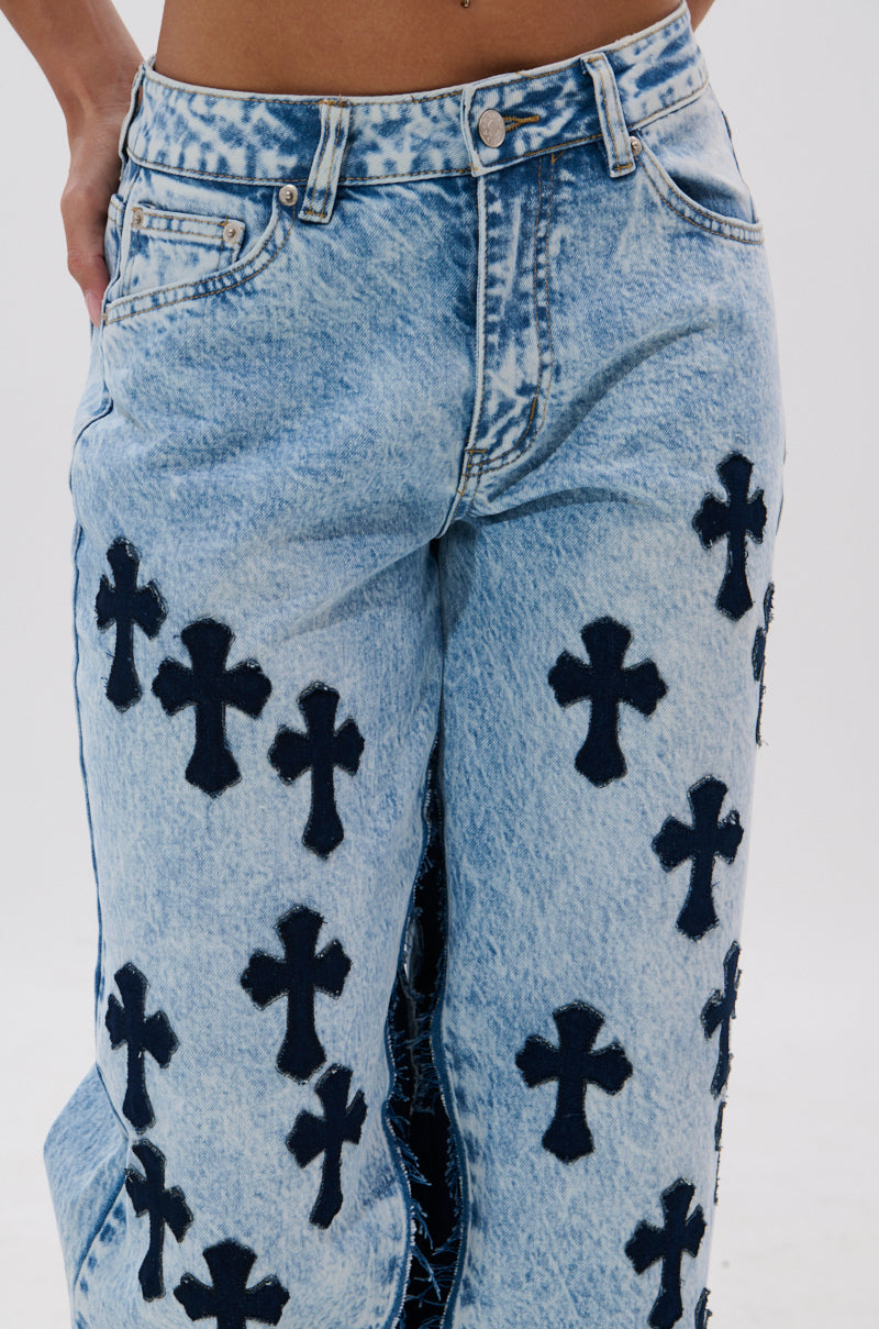 Model wears CROSSED OUT WIDE LEG DENIM—vintage light-wash, high-rise jeans featuring black cross patches on the front and sides. Background is plain and light-colored.