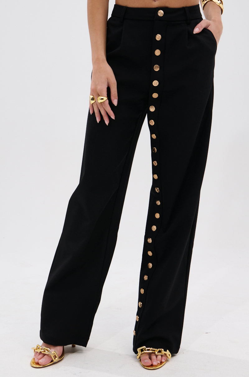Wearing the BUTTONED UP STRAIGHT LEG PANT featuring metallic gold buttons, paired with gold sandals and jewelry, a person stands against a plain light background. 1