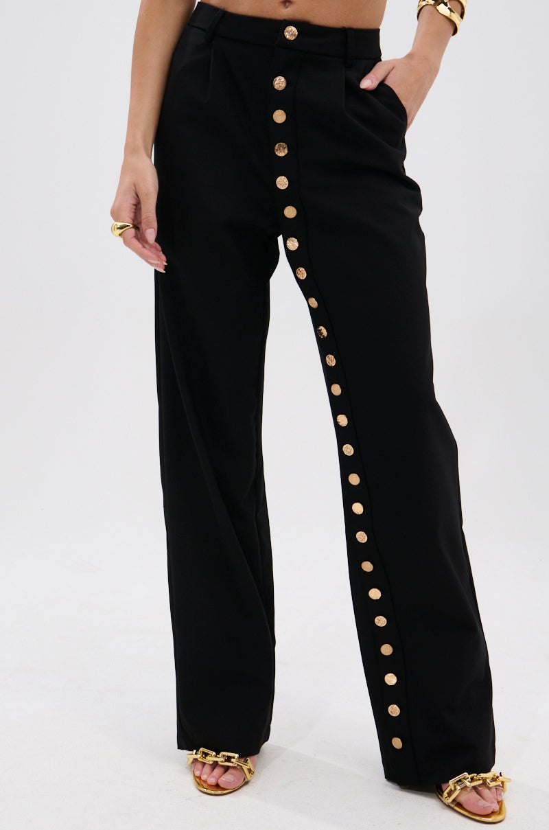 A person wears the BUTTONED UP STRAIGHT LEG PANT—high-waisted black pants with metallic gold buttons—styled with gold jewelry and open-toed gold chain heels. The background is plain white. 2