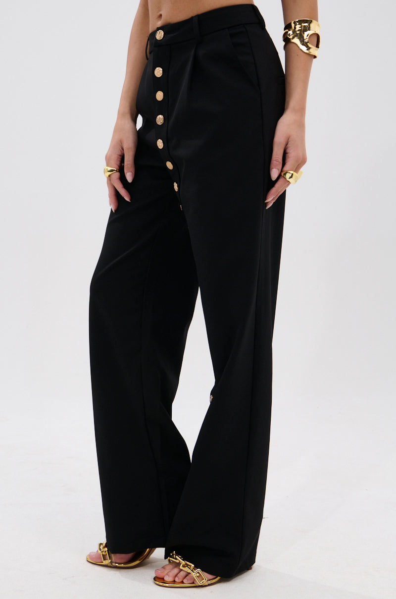 A person stands against a white background wearing the BUTTONED UP STRAIGHT LEG PANT, featuring high-waisted, straight-leg black design with metallic gold buttons, paired with gold sandals and matching gold jewelry. 3