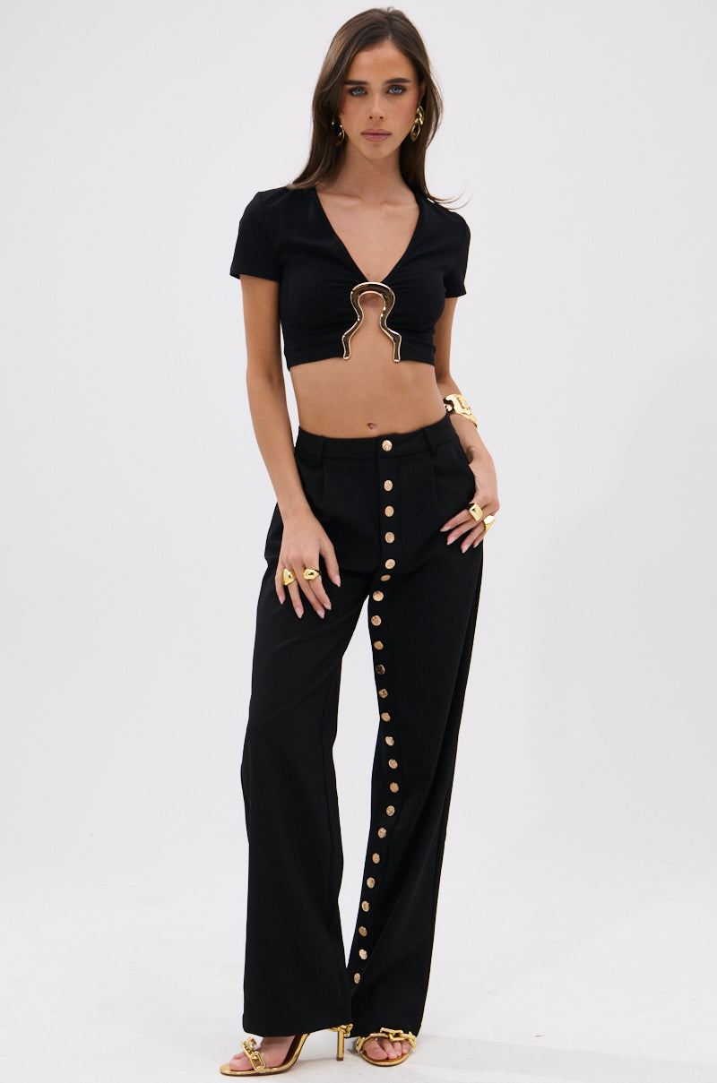 A woman poses in BUTTONED UP STRAIGHT LEG PANT—black, high-waisted with large metallic gold buttons down one leg—paired with a black keyhole crop top, gold jewelry, and heels against a white background. 5