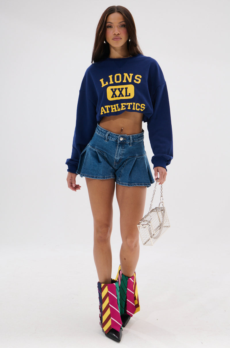 A woman stands against a white background wearing a blue "Lions XXL Athletics" cropped sweatshirt, the NICOLETTE DENIM SHORT, colorful striped boots, and holding a small textured handbag. 0