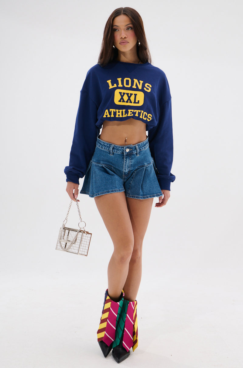 A woman wearing a blue "Lions XXL Athletics" crop sweatshirt, NICOLETTE DENIM SHORT mid-rise shorts with a pleated hem, colorful striped knee-high boots, and holding a small chain-link handbag stands against a plain white background. 4