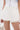 A person wears the GUESS WHO MINI SKORT, a white pleated mini skort with built-in shorts and a high-rise waist, paired with a white corset top featuring button details and a white textured handbag for a chic, modern look.