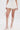 Dressed in the GUESS WHO MINI SKORT, a person stands against a plain white background, paired with a small white handbag and knee-high white gladiator sandals featuring multiple buckled straps and block heels.