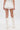 A person faces away, wearing the GUESS WHO MINI SKORT with a pleated overlay, knee-high white cutout boots with bows, and holding a small white handbag against a plain white background.