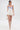 A woman in a strapless blue and white corset top, the GUESS WHO MINI SKORT, knee-high white gladiator heels, a pearl necklace, and holding a white handbag walks confidently against a plain white background.
